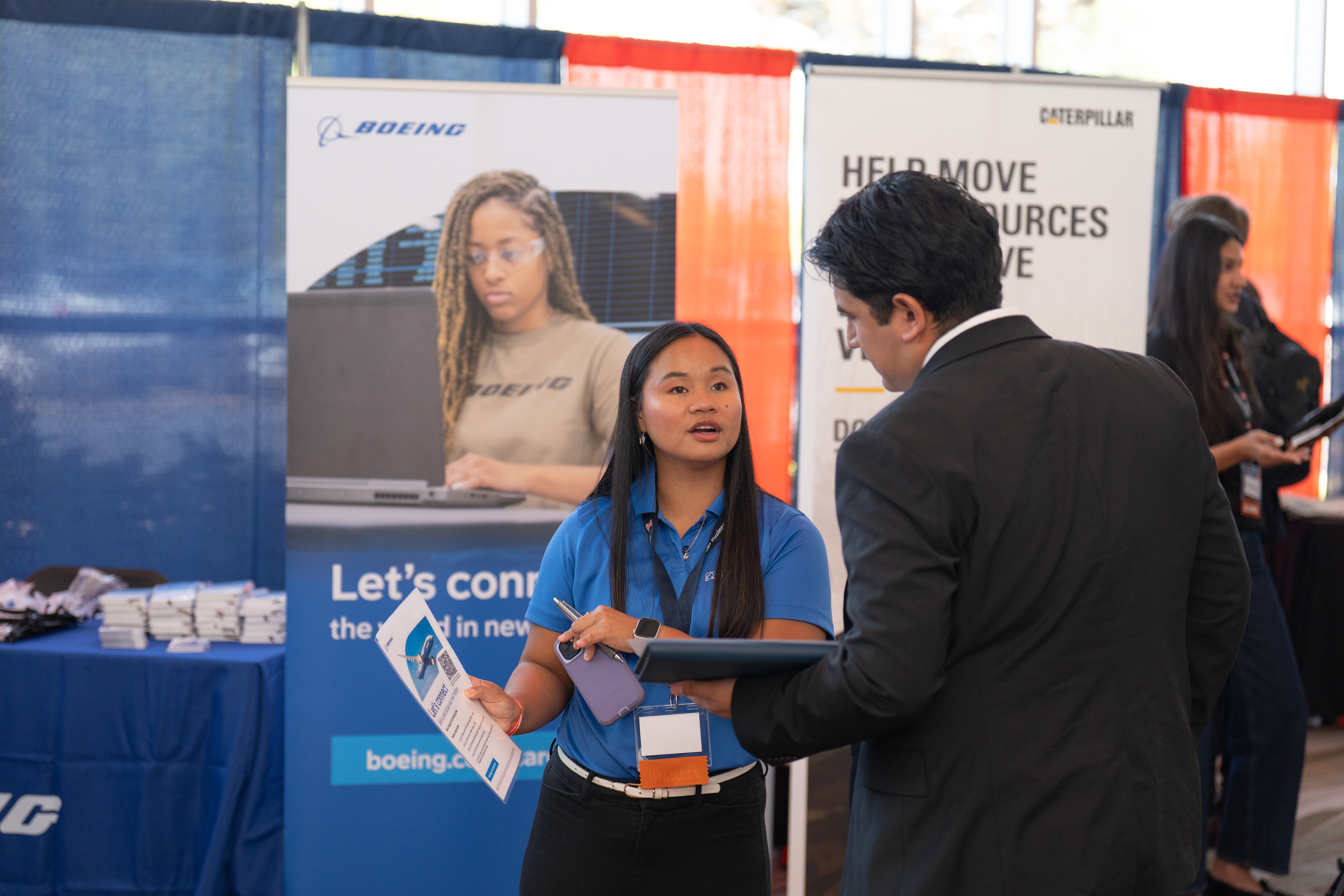 A Femal Boeing Airlines Employee Standing in front of booth. Talking to student.