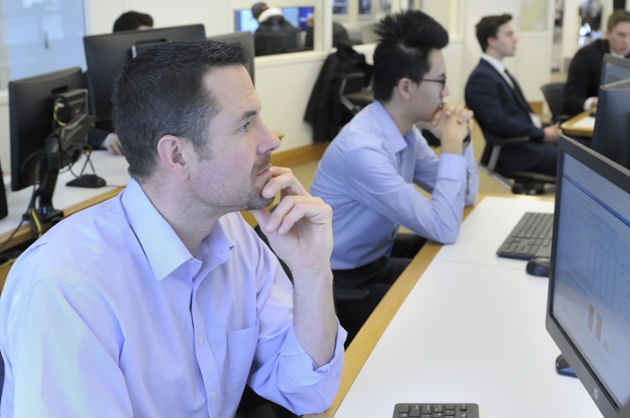 A row of students sit at a computer lab while listening to a speaker