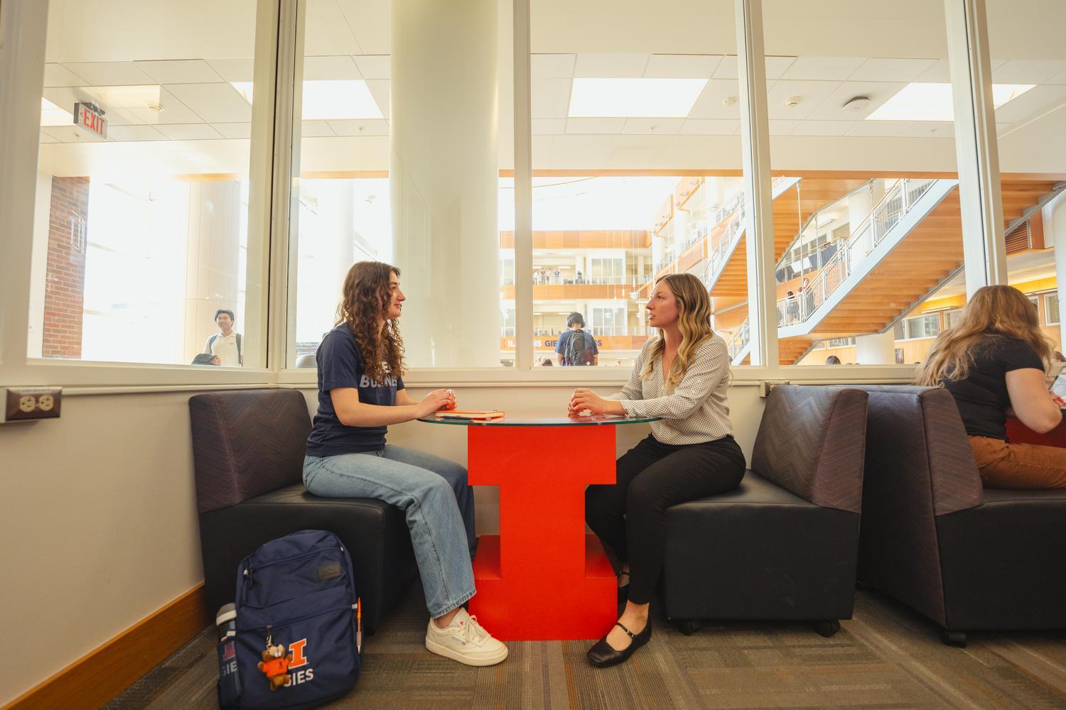 Student in blue shirt and jeans sitting at a table across from an academic advisor in white blouse and black pants.